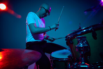 Focused bald drummer in white shirt playing in pink-purple stage lighting against gradient studio...