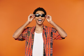 cheerful young african american man in vibrant attire with sunglasses posing on orange backdrop