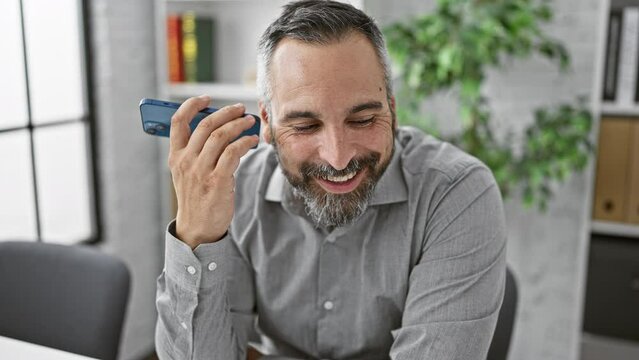 Handsome mature hispanic man with grey beard smiling and talking on smartphone in modern office interior.