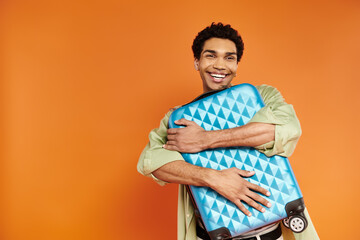 handsome joyful african american man in stylish attire hugging his blue suitcase on orange backdrop