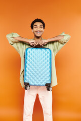 cheerful handsome african american man in casual outfit holding blue suitcase and smiling at camera