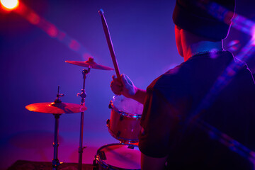 Back view portrait of young artistic drummer performing in pink-purple stage lighting against...