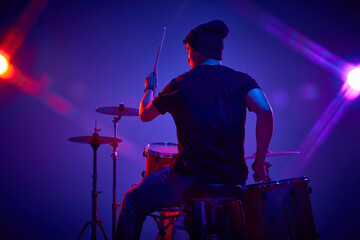 Rear view portrait of young artistic man playing drums in pink-purple stage lighting against...