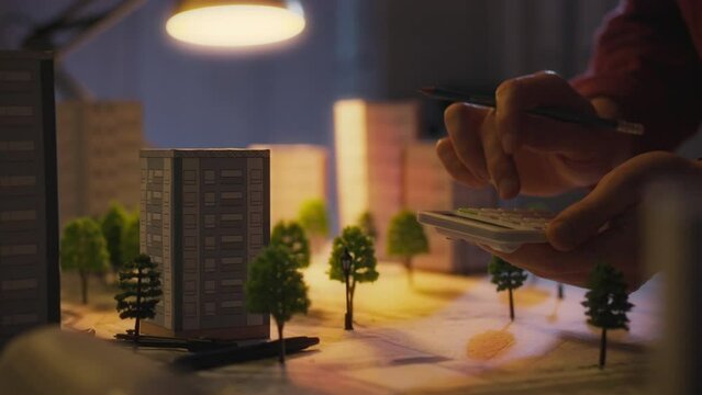 Late in the day, the architect actively uses a calculator to make calculations. Under the light of a desk lamp, an engineer analyzes data needed to design a city of the future