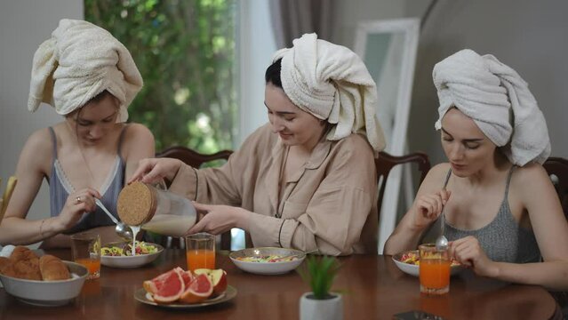 Three girls with towels on their heads are having breakfast sitting at a table in the living room. A girl adds milk from a glass jug to her friend breakfast cereal on a plate. The girls eat and laugh