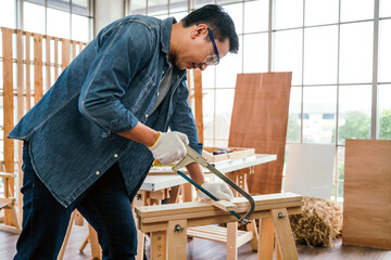 Asian father and son work as a woodworker or carpenter, Father wears safety goggles and saw a wooden plank with hacksaw carefully. Craftsman carpentry working at home workshop studio. Small business
