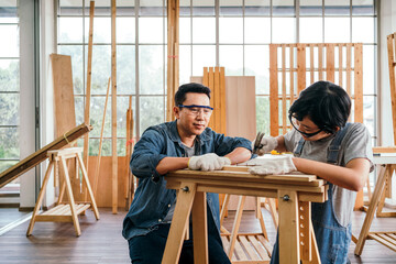 Happy Asian father and son work as a woodworker and carpenter. Father teaching his son to hammer nails on a wooden plank carefully together. carpentry working at a home workshop studio. Small business - 783067355