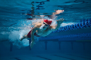 Athletic grace and strength. Young man in cap and goggles, swimmer in motion underwater training in...