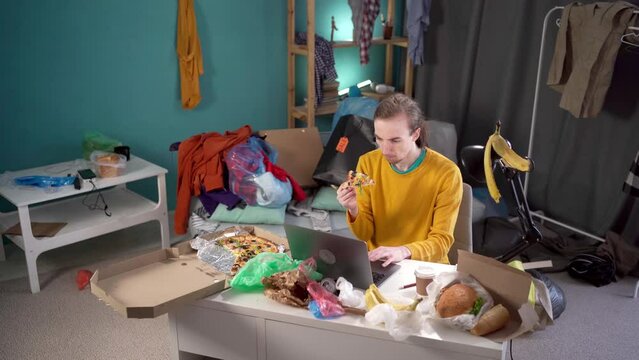 Young man working studying in messy room sitting at the table with food and garbage.