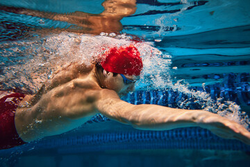 Discipline and focus. Muscular, athletic young man in red can and goggles in motion, swimming in...