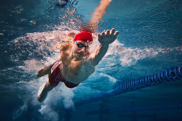 Resilience and strength. Concentrated young man in red cap and goggles in motion, swimming in pool, training. Concept of professional sport, health, endurance, strength, active lifestyle - obrazy, fototapety, plakaty