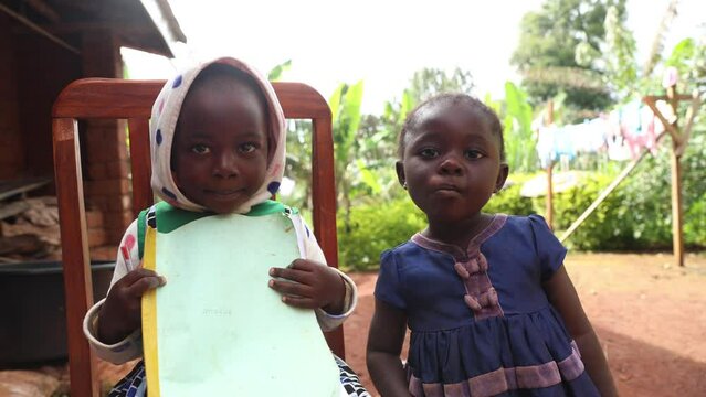 Two young children are standing together in the village, one holding a book, shy African kids