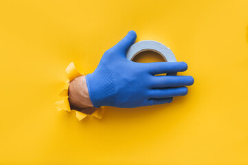 A right man's hand in a blue fabric work glove holds a roll of white double-sided tape. Torn hole in yellow paper. Repair work. Copy space.