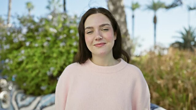A smiling young woman with brown hair, dressed in a pink sweater, enjoying a sunny day in a natural park setting with palm trees in the background.