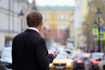 Man in a business suit standing with smartphone on a street on cars background. Using mobile phone...