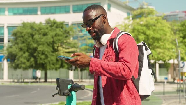 Happy young generation z African Black man tourist traveler wearing headphones and backpack having fun riding electric bike scooter exploring city street outdoors on travel trip on sunny day.