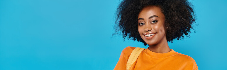 A college girl with a large afro smiles warmly at the camera against a blue backdrop in a studio...