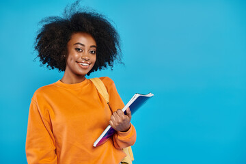 A young African American college girl standing in casual attire, holding a book, smiling warmly at...
