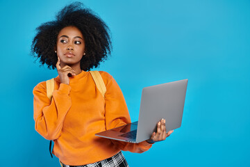 Obraz na płótnie Canvas African American college girl in casual attire standing with laptop in front of blue background.
