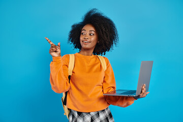 An African American college girl in casual attire holding a laptop, pointing to the side in a studio with a blue backdrop. - obrazy, fototapety, plakaty