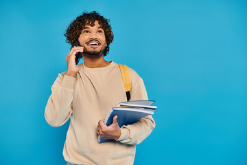 An Indian student in casual attire stands against a blue backdrop, holding a folder and talking on...