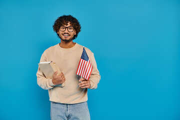 A man proudly holds a book and an American flag, standing against a blue backdrop in a studio.
