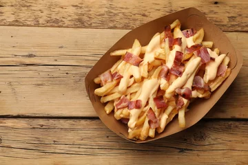 Poster Tasty potato fries, cheese sauce and bacon in paper container on wooden table, top view. Space for text © New Africa