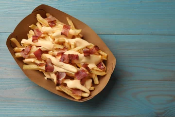 Tuinposter Tasty potato fries, cheese sauce and bacon in paper container on light blue wooden table, top view. Space for text © New Africa