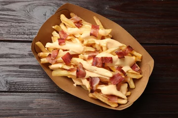 Tuinposter Tasty potato fries, cheese sauce and bacon in paper container on wooden table, top view © New Africa