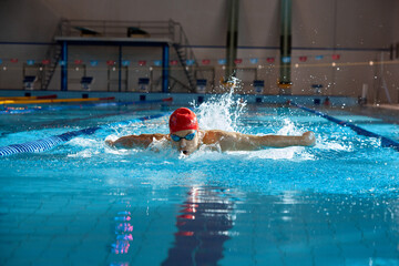 Competitive, muscular young man, swimmer in red cap performs butterfly stroke in pool, training...