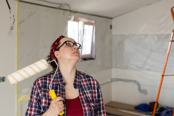 A woman in glasses and a plaid shirt with a roller in her hands looks up while examining a surface for painting. High quality photo