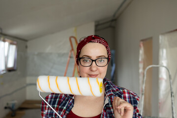 A smiling girl in glasses with a roller in her hands is preparing to start painting the walls. High quality photo