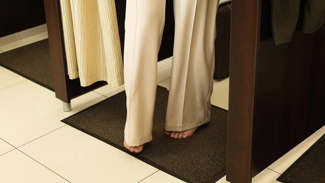 Close-up of woman's legs getting dressed, trying on beige trousers in store changing room. Shopping for casual clothes in boutique.