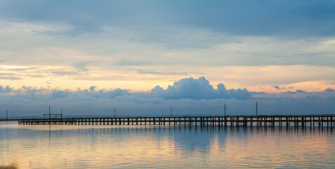 The sky is cloudy and the water is calm with a pier in the mid-ground