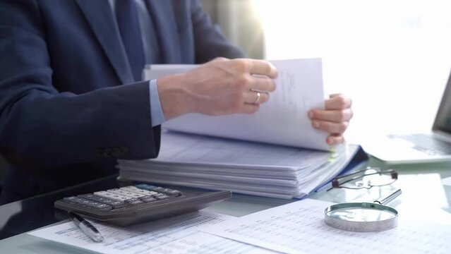 Businessman using a calculator and magnifying glass while analyzing financial documents at desk in fair modern office. Audit and taxes in business