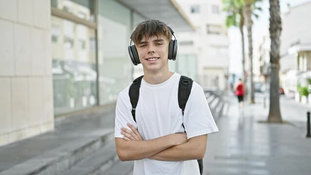 A smiling young caucasian male teenager with headphones and a backpack stands confidently with his arms crossed on a city street.