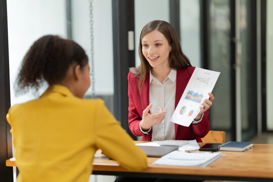 Two businesswomen review financial charts and reports in a modern office, collaborating, discussing strategy and growth while smiling and analyzing data during a meeting - Powered by Adobe