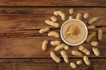 Delicious nut butter in bowl and peanuts on wooden table, top view. Space for text