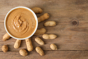 Delicious nut butter in bowl and peanuts on wooden table, top view. Space for text