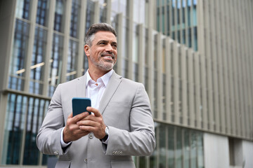 Happy middle aged business man ceo entrepreneur investor standing outside office using mobile cell phone looking away. Mature businessman professional executive holding smartphone working on cellphone - 783153566
