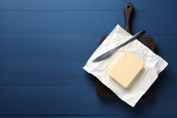 Block of tasty butter in open foil packaging and knife on blue wooden table, top view. Space for...