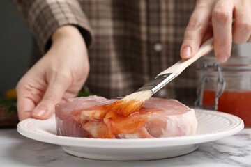 Woman spreading marinade onto raw meat with basting brush at white marble table, closeup