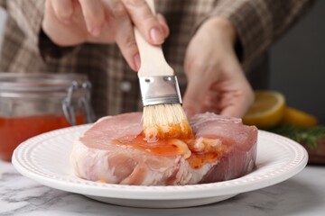 Woman spreading marinade onto raw meat with basting brush at white marble table, closeup