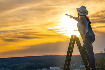 a woman in a work pants stands on a wooden ladder and paints the sunset