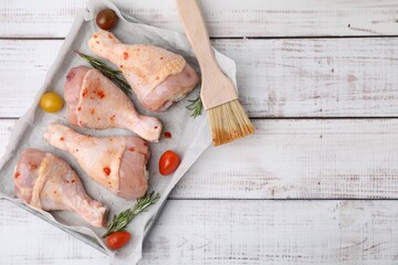 Raw marinated chicken drumsticks, rosemary, tomatoes and basting brush on white wooden table, flat...