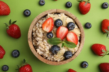 Tasty oatmeal with strawberries, blueberries and almond petals in bowl surrounded by fresh berries on green background, flat lay