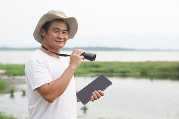 Handsome Asian man ecologist is surveying nature at the lake, holds binoculars and smart...