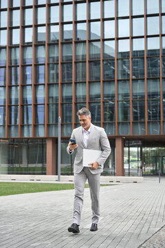 Middle aged businessman using phone walking on city street, busy older business man leader investor, mature male executive in suit holding laptop going near office building, full body vertical photo.