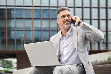 Busy mature business man merchant making phone call using laptop sitting outside office. Happy middle aged businessman manager wearing suit talking on cellphone working on computer consulting client. - 783163328