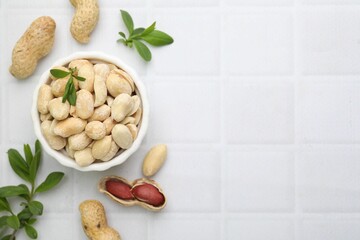 Fresh peeled peanuts in bowl and leaves on white tiled table, flat lay. Space for text